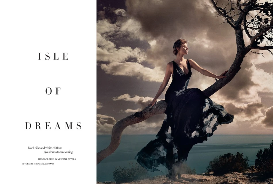 Elegant woman in black dress sits on tree branch against dramatic cloudy sky and ocean backdrop in a fashion editorial style photoshoot.
