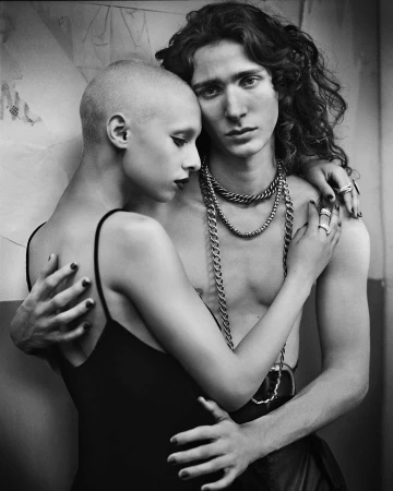 Black and white image of a shirtless man with long curly hair embracing a bald woman in a black dress, both wearing rings.