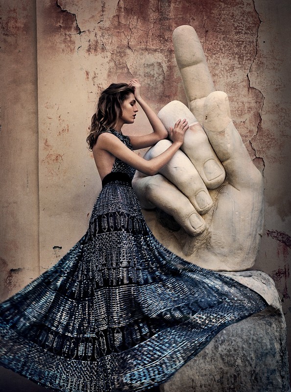 Elegant woman in flowing blue dress posing next to a large stone hand sculpture against a weathered wall backdrop.