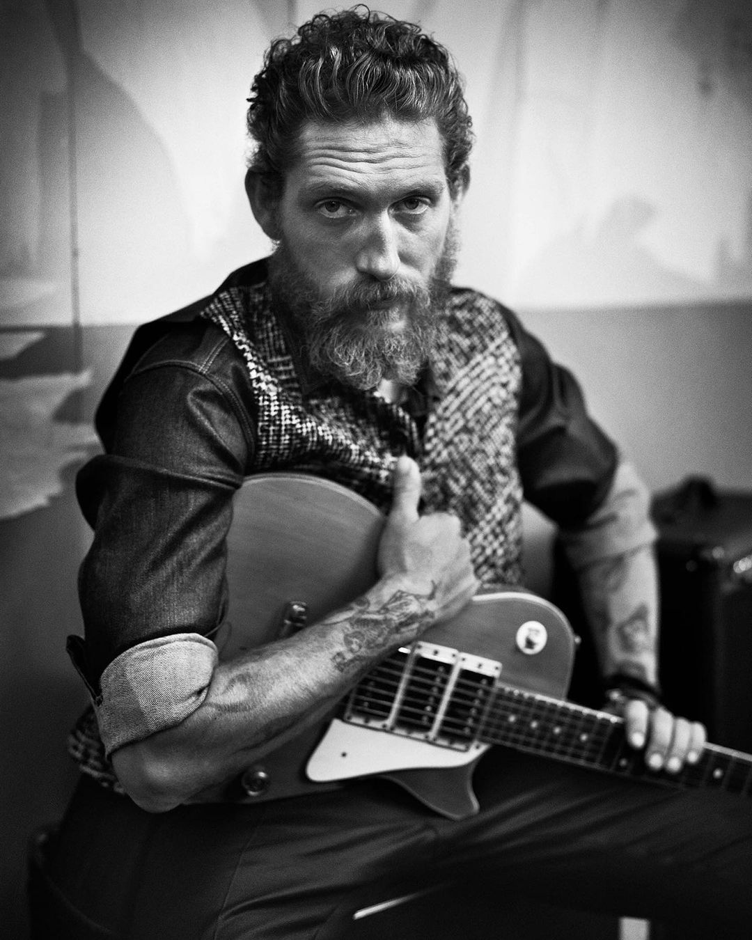 Black and white portrait of a man with a beard holding an electric guitar, looking directly at the camera with a serious expression.