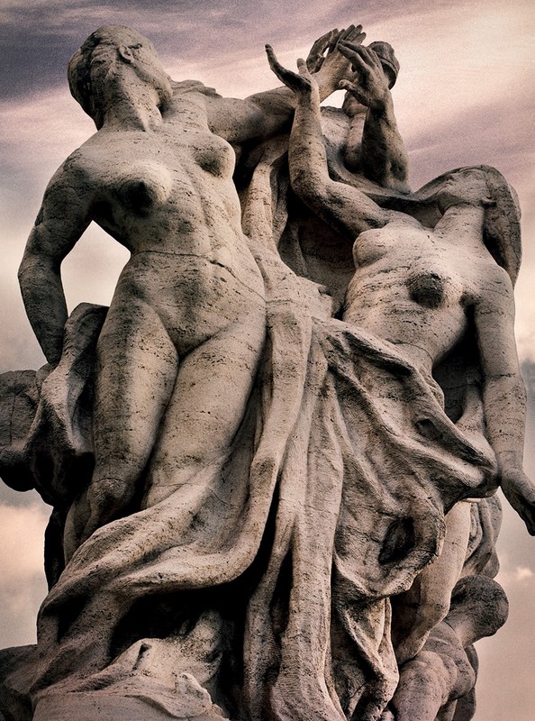 Stone sculpture of three nude figures, one figure raising hands in the air, set against an overcast sky. Fine art nude.