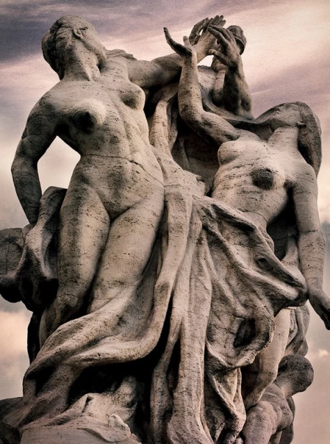 Stone sculpture of three nude figures, one figure raising hands in the air, set against an overcast sky. Fine art nude.