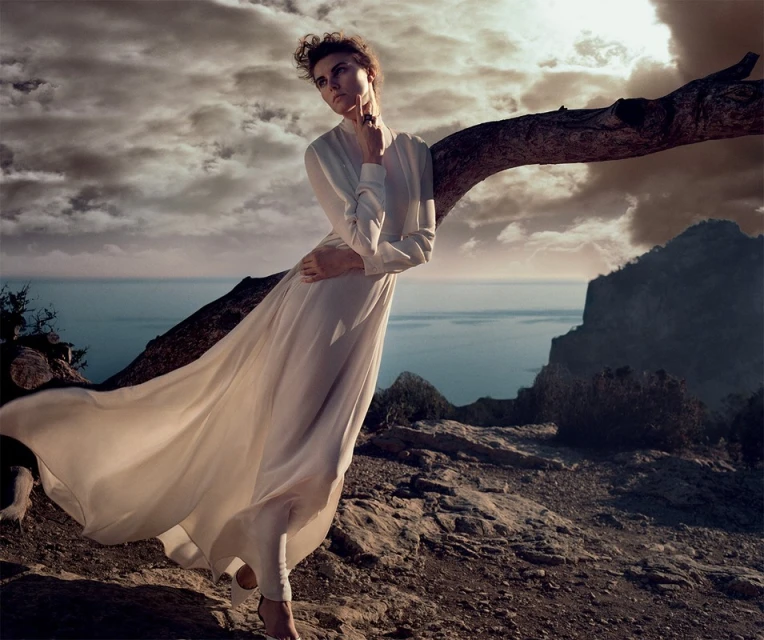 A woman in a white dress is leaning against a tree branch, with the ocean and cloudy sky visible in the background, fashion photography.