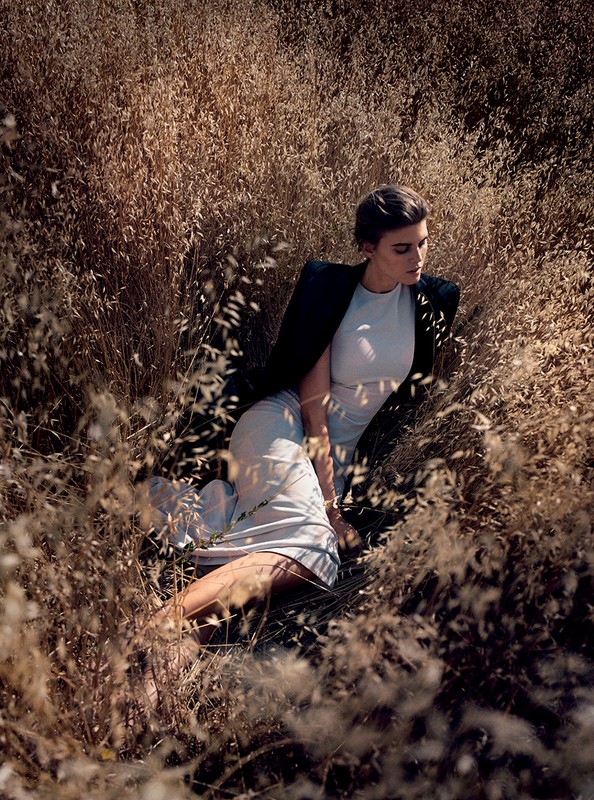 Woman in a white dress and black jacket lies in a field of tall, golden grass with a shallow depth of field in a natural, outdoor setting.