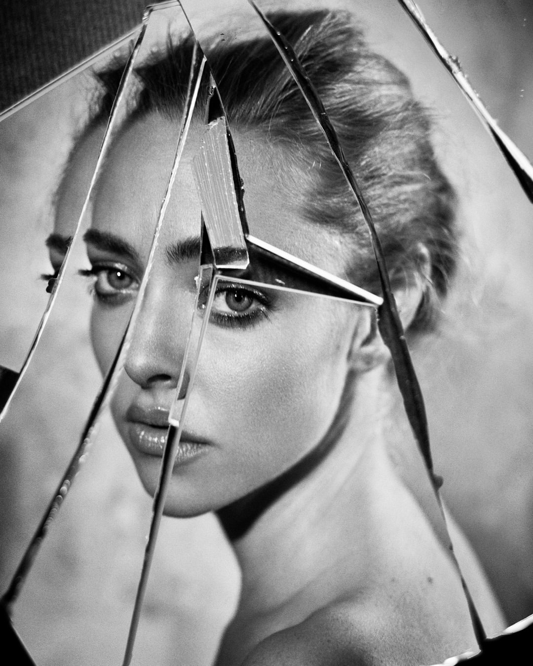 Striking black and white portrait of a woman with clear eyes, partially obscured by shards of a broken mirror, creating fractured reflections.