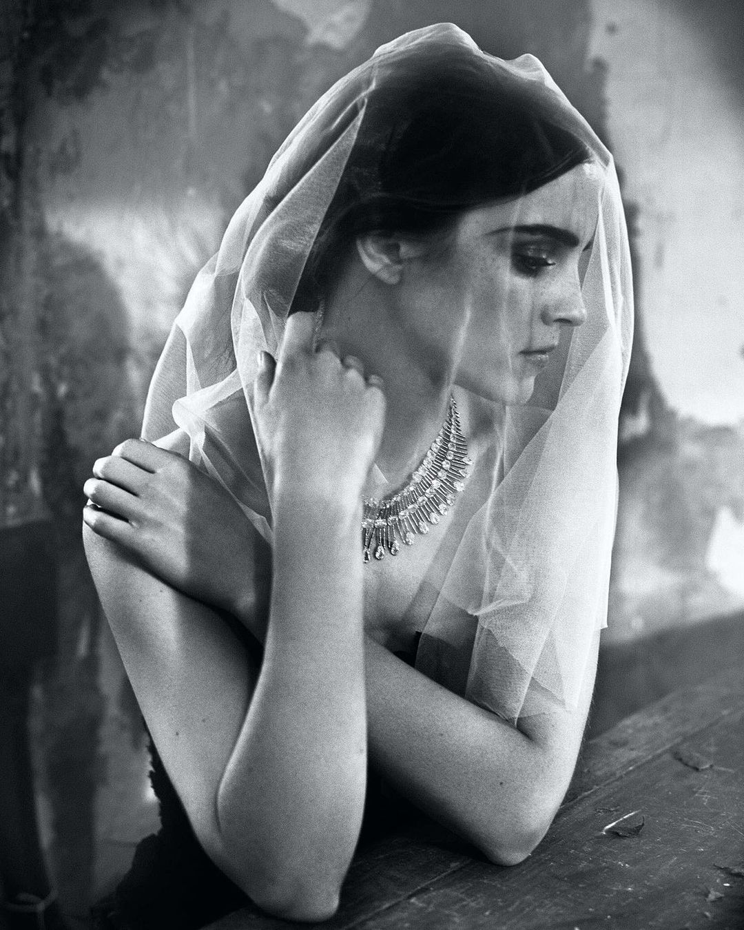 Monochrome portrait of a female model with her arms crossed, wearing a sheer veil and a jeweled necklace, against a textured backdrop.