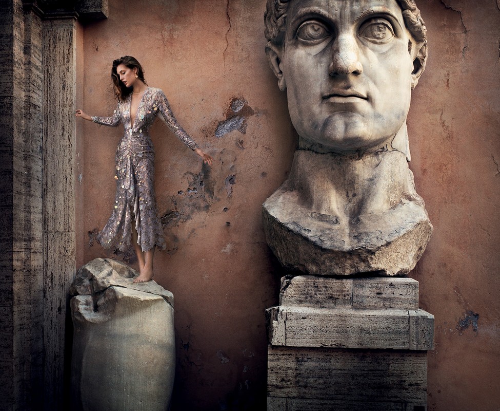 Fashion shot of a woman in a dress on a marble column, next to a marble bust against a textured wall, creating a vintage aesthetic.