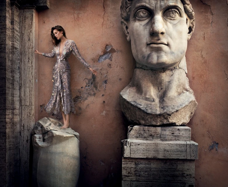 Fashion shot of a woman in a dress on a marble column, next to a marble bust against a textured wall, creating a vintage aesthetic.