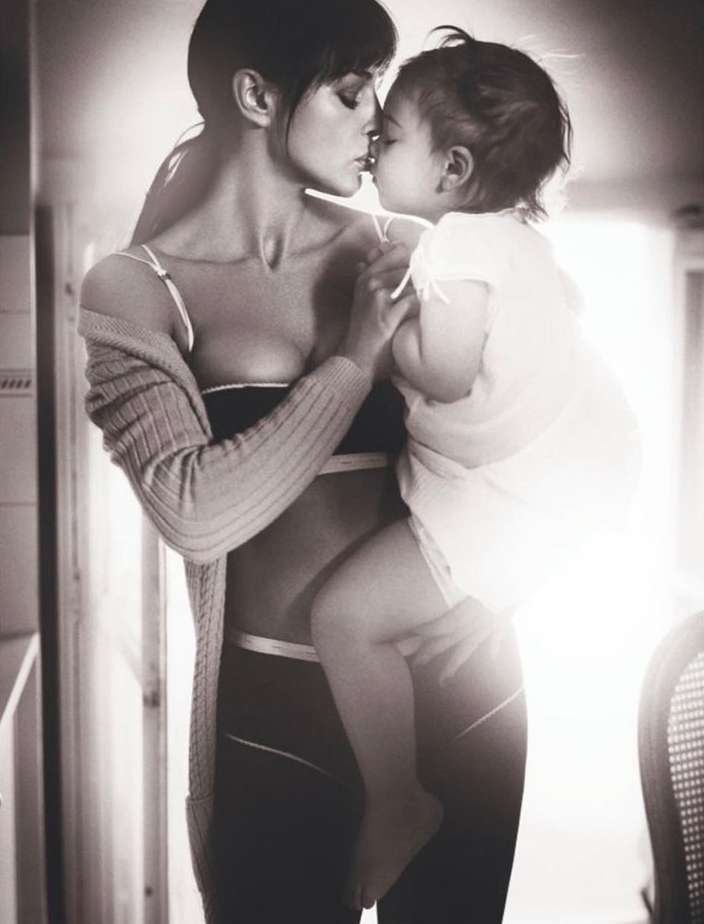 Monochrome portrait of a woman holding a toddler, faces close together in kissing gesture, backlit indoor setting with minimal background.