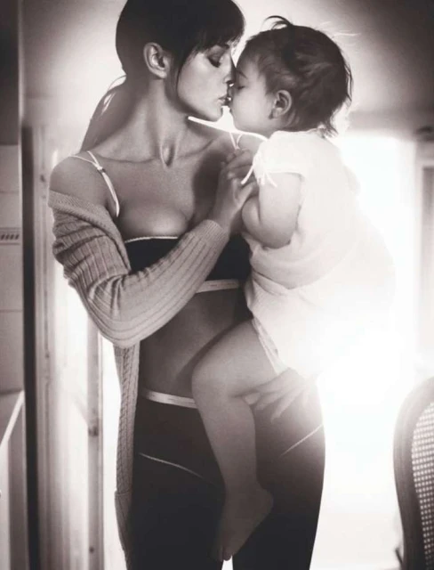 Monochrome portrait of a woman holding a toddler, faces close together in kissing gesture, backlit indoor setting with minimal background.