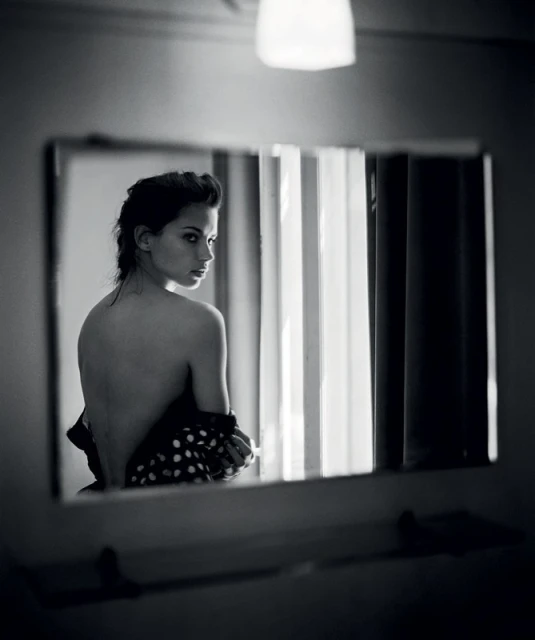 Black and white portrait of a woman looking over her shoulder in a mirror. Polka dot fabric, light fixture, and neutral background.