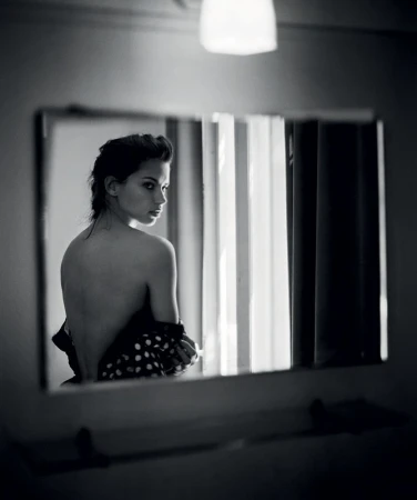 Black and white portrait of a woman looking over her shoulder in a mirror. Polka dot fabric, light fixture, and neutral background.