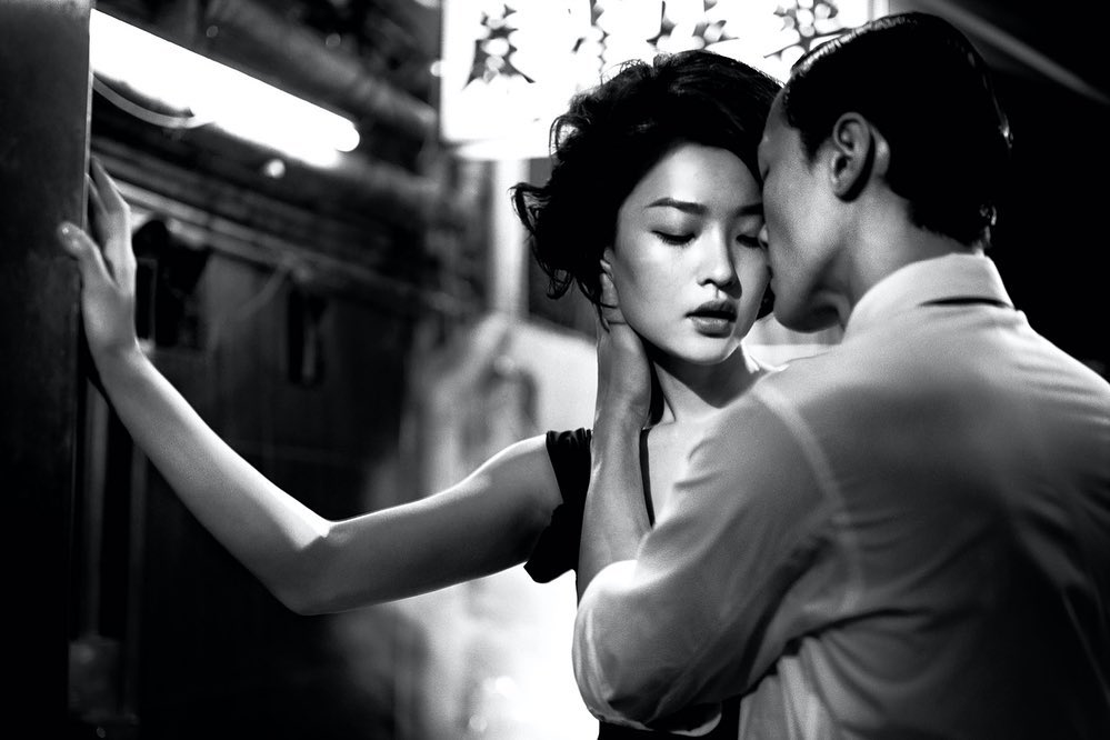 A couple embraces closely in a black and white photo. The woman leans against a wall as the man appears to be whispering in her ear.