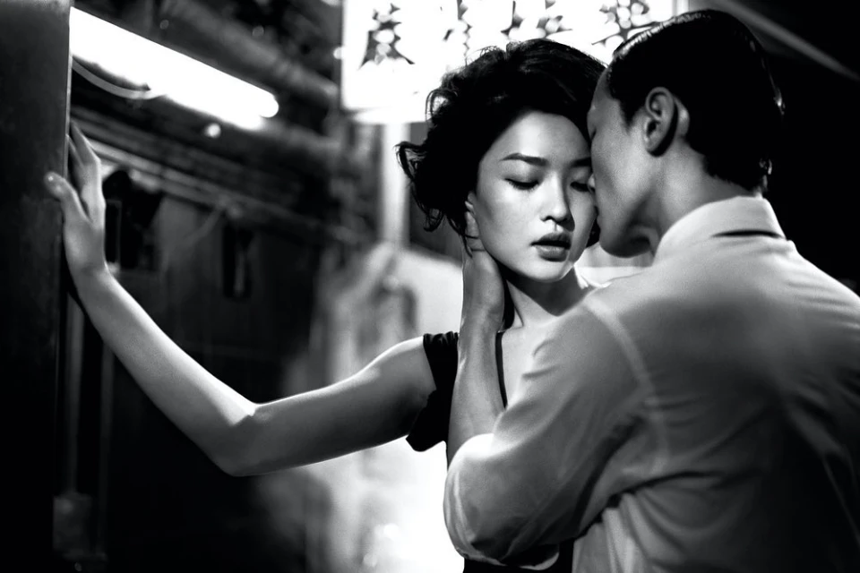 A couple embraces closely in a black and white photo. The woman leans against a wall as the man appears to be whispering in her ear.