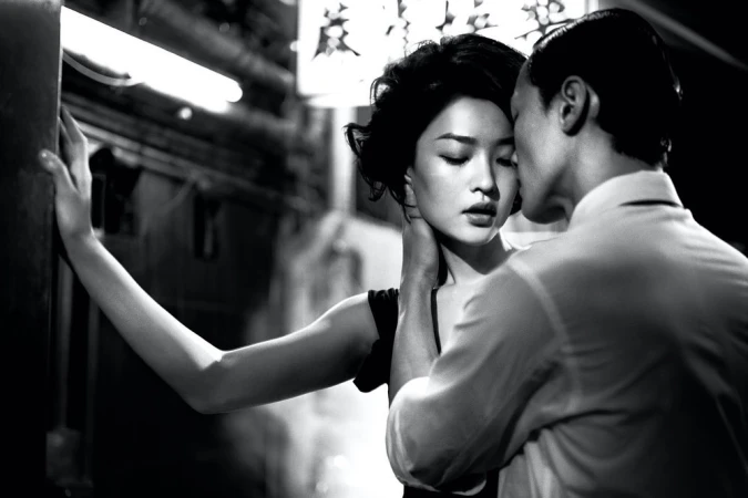 A couple embraces closely in a black and white photo. The woman leans against a wall as the man appears to be whispering in her ear.