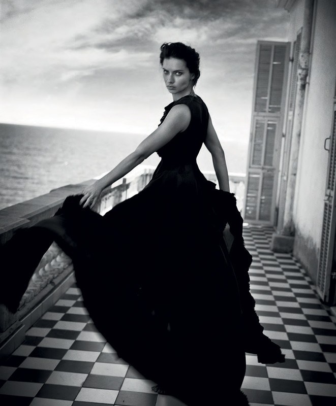 Monochrome portrait of a woman in a black dress posing on a tiled balcony with a sea view, gazing at the camera in a fashion shot.