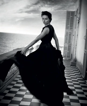 Monochrome portrait of a woman in a black dress posing on a tiled balcony with a sea view, gazing at the camera in a fashion shot.