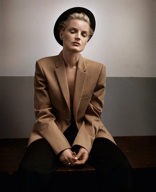 A woman wearing a camel blazer and black hat sitting down against a studio wall, photographed by Vincent Peters.