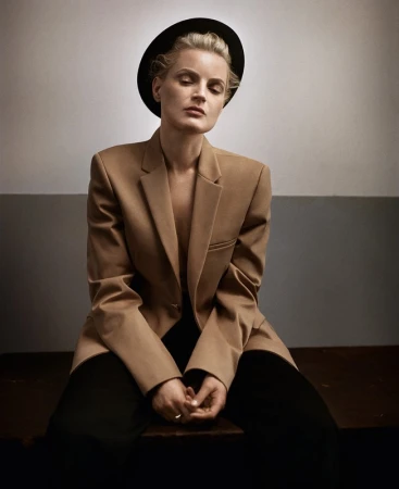 A woman wearing a camel blazer and black hat sitting down against a studio wall, photographed by Vincent Peters.