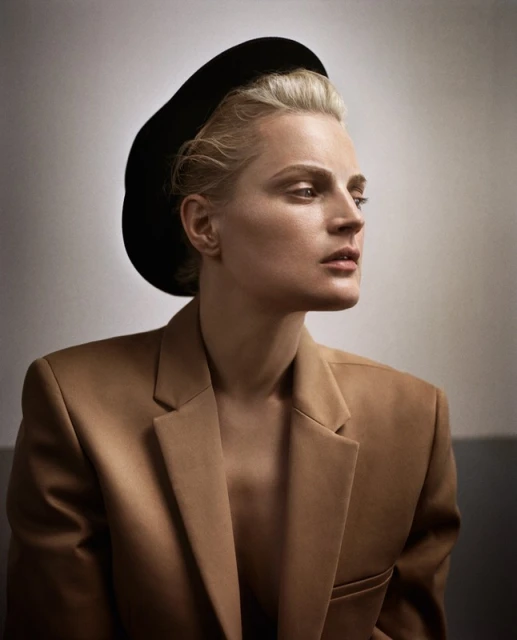 Portrait of a blonde adult woman in a tan jacket and black hat, looking off to the side in a studio setting with neutral background.