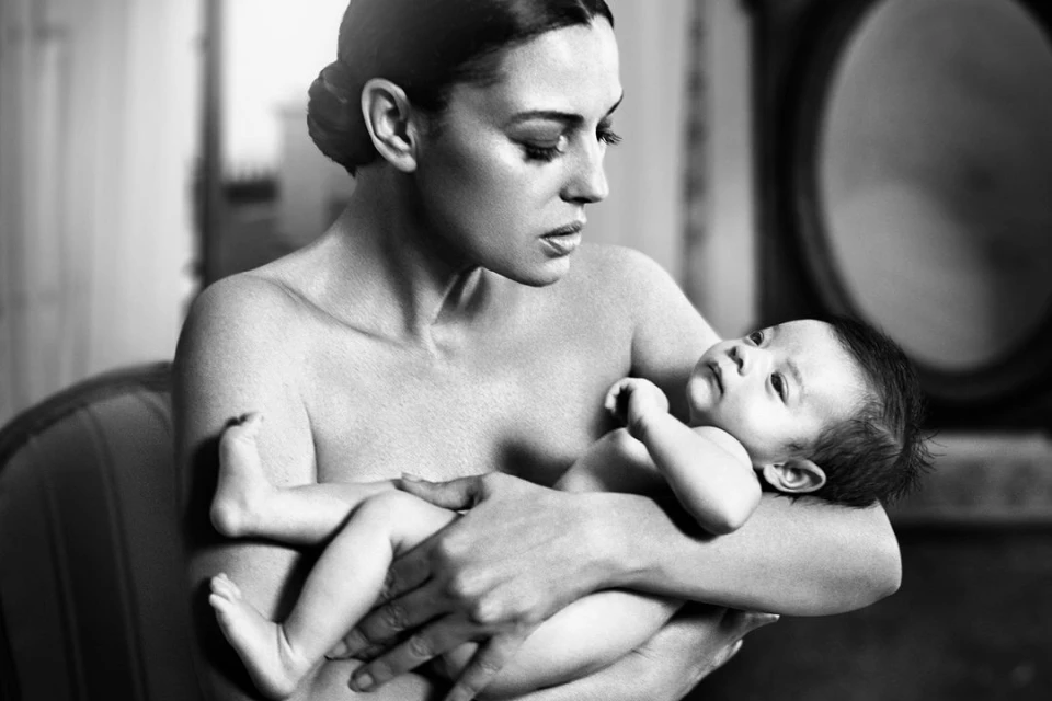 Black and white portrait of adult woman holding baby indoors, showcasing a tender moment with soft lighting and neutral background.