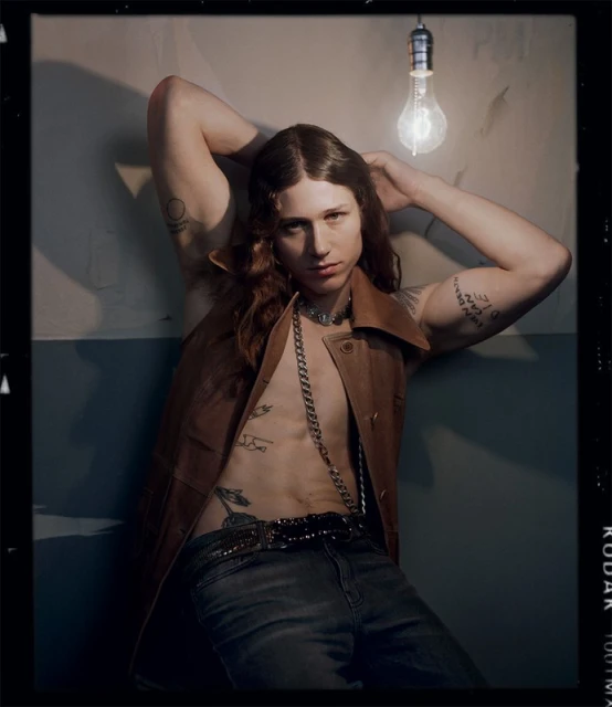 Portrait of a young adult male with long hair and tattoos, wearing a vest and jeans, leaning against a wall with a bare lightbulb above.