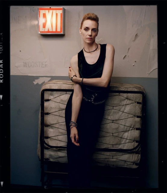 A woman in a black dress poses indoors, leaning against a bed frame, with an orange EXIT sign and worn wall in the background.