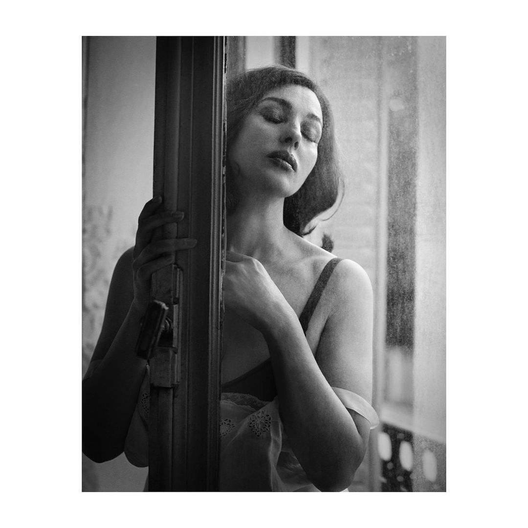 Black and white portrait of an adult female with eyes closed, positioned by a window and door frame, with soft lighting.
