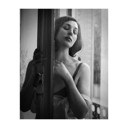 Black and white portrait of an adult female with eyes closed, positioned by a window and door frame, with soft lighting.