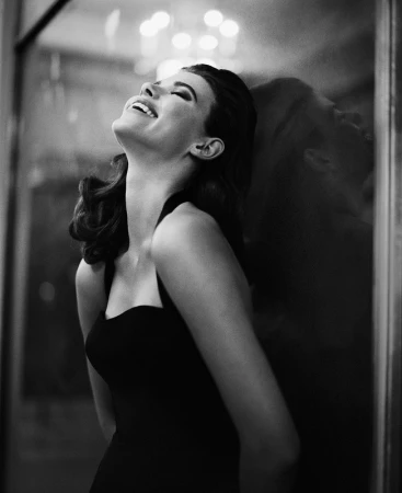 Monochrome portrait of a smiling woman in a black halter dress leaning against a dark wall by Vincent Peters.