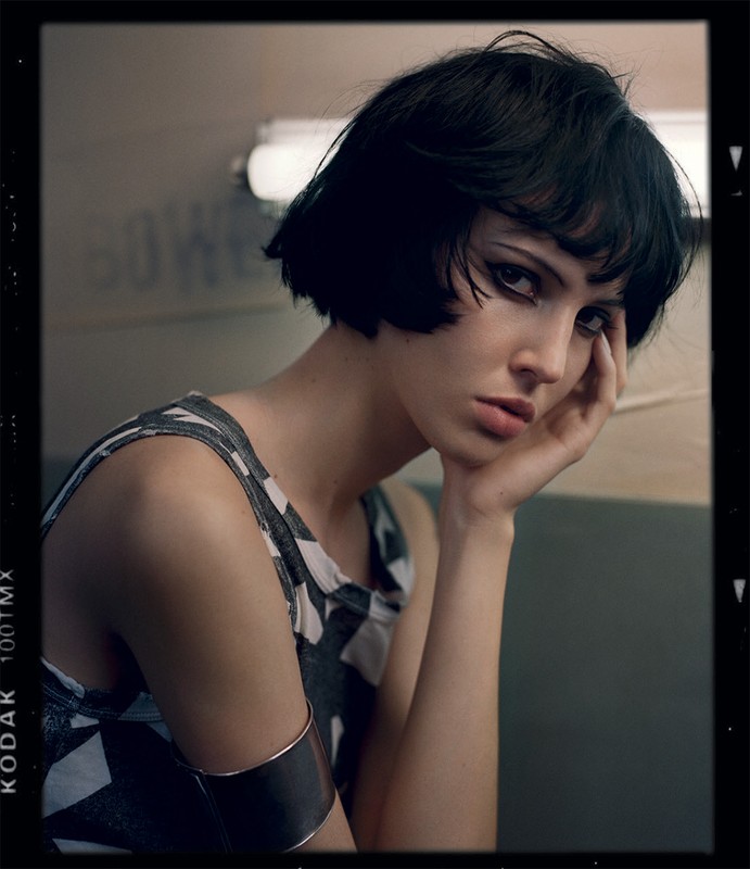 A woman with dark hair and dramatic eye makeup poses indoors wearing a patterned top and cuff bracelet, portrait in a filmic style.