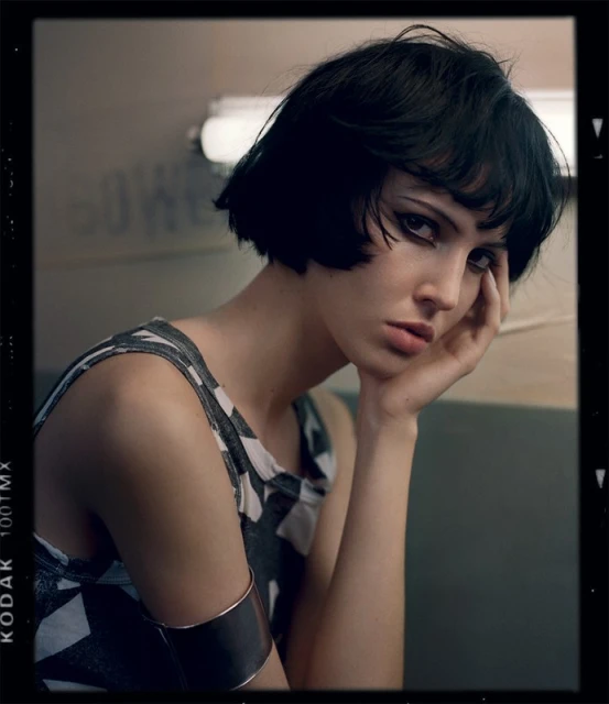 A woman with dark hair and dramatic eye makeup poses indoors wearing a patterned top and cuff bracelet, portrait in a filmic style.
