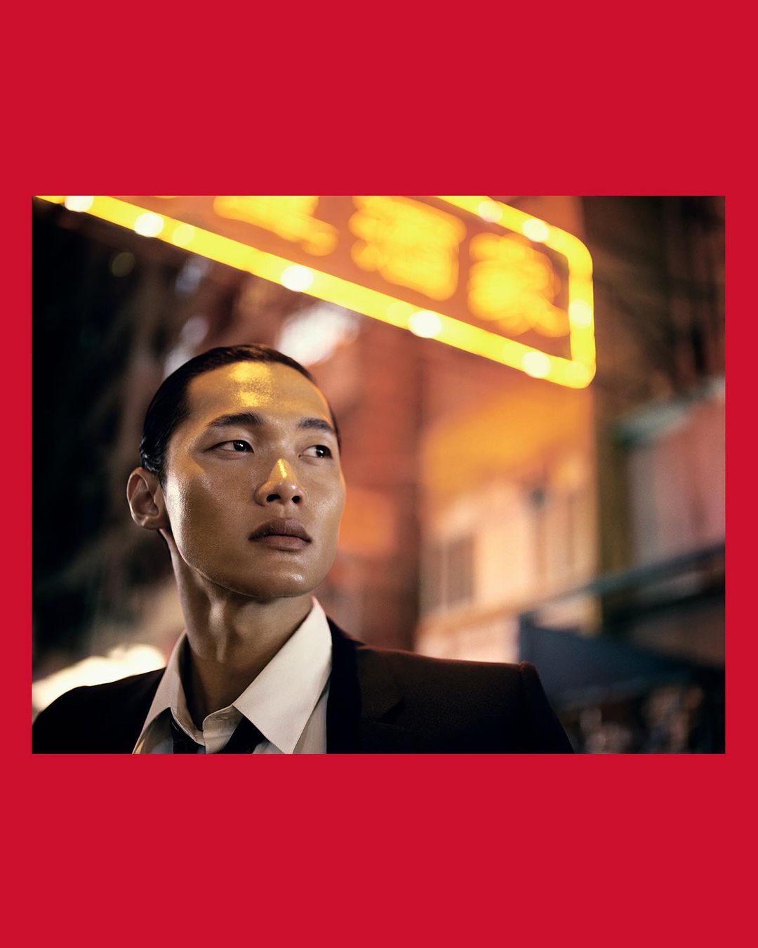 Portrait of an Asian man in a dark suit and white shirt with a neon sign in the blurred background, framed by red borders.