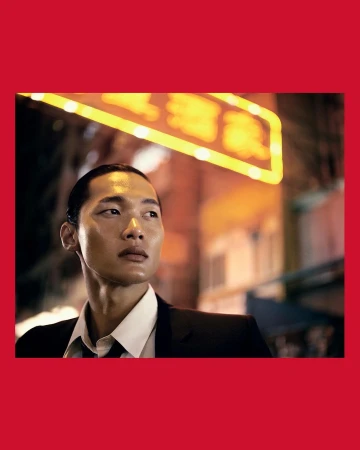 Portrait of an Asian man in a dark suit and white shirt with a neon sign in the blurred background, framed by red borders.