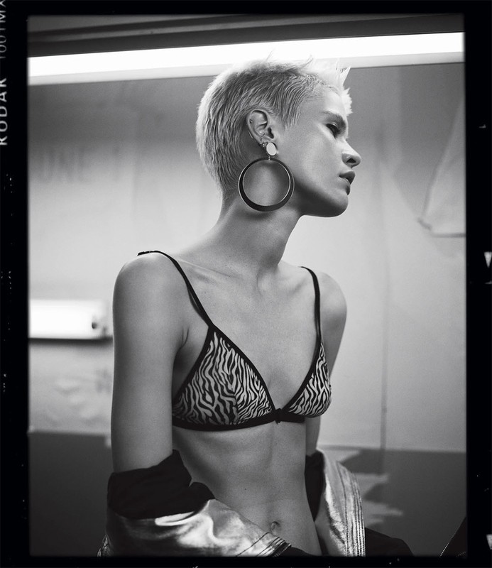 Monochrome portrait of a slender adult woman with short blonde hair, wearing a zebra-print bikini top, jewelry, and a jacket, looking away.