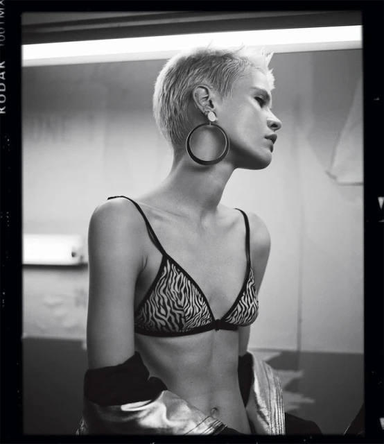 Monochrome portrait of a slender adult woman with short blonde hair, wearing a zebra-print bikini top, jewelry, and a jacket, looking away.