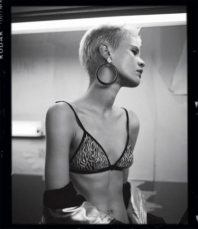 Monochrome portrait of a slender adult woman with short blonde hair, wearing a zebra-print bikini top, jewelry, and a jacket, looking away.