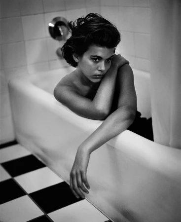 Black and white portrait of a woman in a bathtub, looking thoughtful with her arm resting on the edge and tile floor visible.