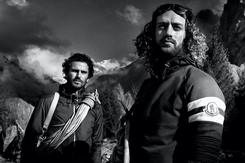 Two adult males stand outdoors in mountaineering attire before a scenic mountain backdrop on a sunny day in a monochrome photograph.