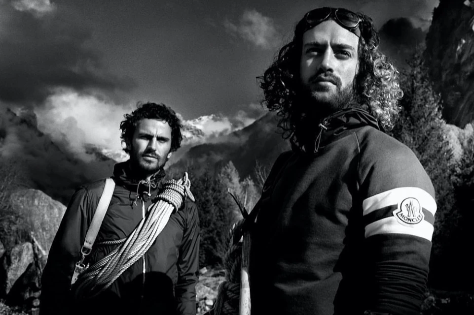 Two adult males stand outdoors in mountaineering attire before a scenic mountain backdrop on a sunny day in a monochrome photograph.