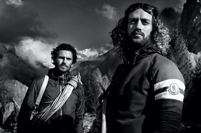 Two adult males stand outdoors in mountaineering attire before a scenic mountain backdrop on a sunny day in a monochrome photograph.