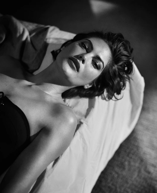 Black and white portrait of an adult woman lying on a surface in dim light, looking at the camera, wearing a strapless top.