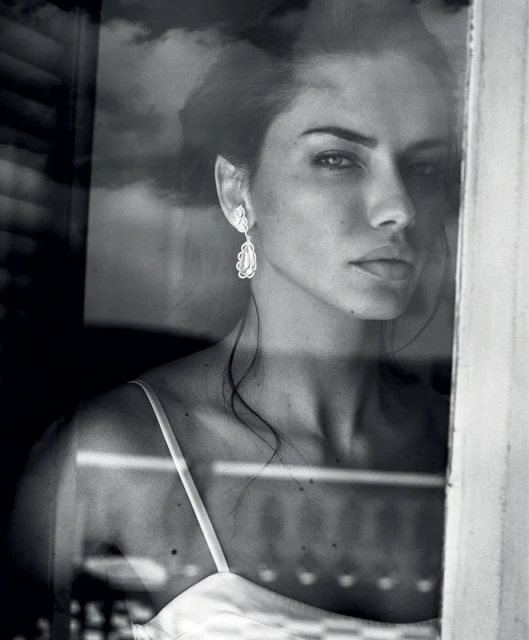 Monochrome portrait of a woman looking through a glass window, showcasing her earrings and shoulder straps in soft lighting.