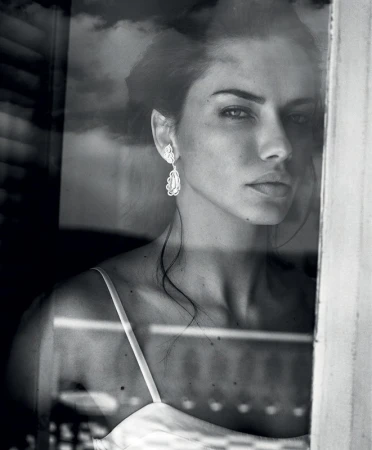 Monochrome portrait of a woman looking through a glass window, showcasing her earrings and shoulder straps in soft lighting.