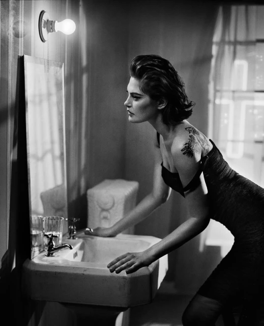 Monochrome portrait of a woman with a tattoo leaning on a bathroom sink under a bare lightbulb, looking at her reflection in the mirror.