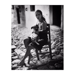 Monochrome portrait of a woman seated on a wooden chair outdoors on cobblestones, holding a small puppy in her arms.