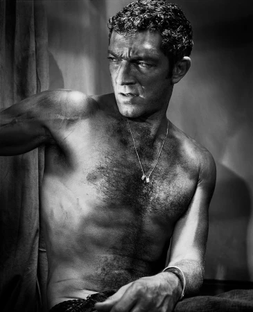 Monochrome portrait of Vincent Cassel shirtless, wearing a thin chain necklace and dark trousers against a plain background.