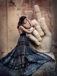 Woman in patterned dark dress leaning against a large sculpted stone hand sculpture