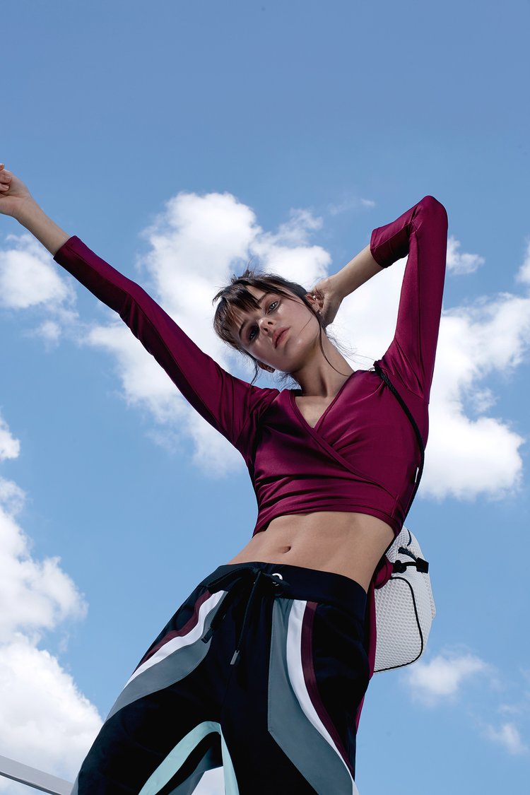 A woman in athletic wear poses with arms raised against a bright blue sky with scattered clouds, showcasing a dynamic outdoor fashion shot.