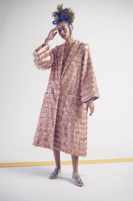 Elegant woman wearing a fringed peach coat and silver shoes in a fashion studio on a white backdrop with a yellow floor line.